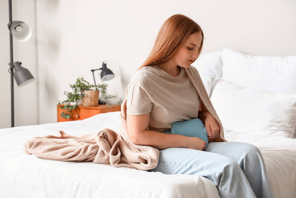 Young woman with hot water bottle having menstrual cramps in bedroom - Foto, Imagen