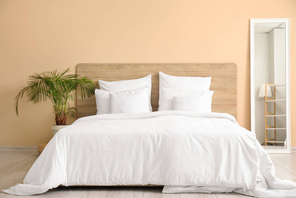 Cómoda cama con almohadas blancas, espejo y planta de interior en el dormitorio - Foto, Imagen