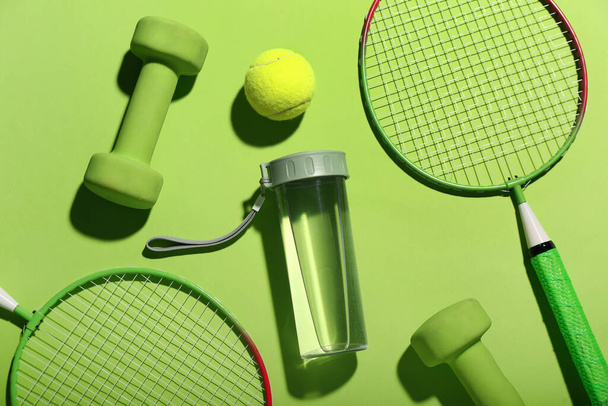Composição com garrafa de água, halteres, raquetes de tênis e bola sobre fundo verde - Foto, Imagem