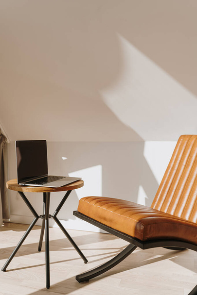Ästhetischer Luxus moderner Home-Office-Arbeitsplatz mit Laptop-Computer, orangefarbenem Ledersessel und Couchtisch. Sonnenlicht wirft Schatten auf die Wand. Lounge-Raum zum Entspannen und Chillen - Foto, Bild
