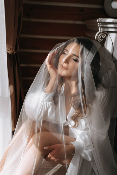 Невеста готовится к свадьбе. Мечтательная девушка в халате с длинными темными волосами возле окна под вуалью - Фото, изображение
