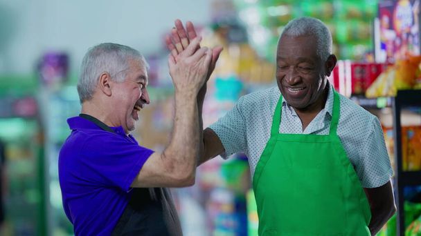 Szczęśliwie zróżnicowani starsi pracownicy świętują sukces przybijając piątkę w nawie supermarketów. Menedżer rasy kaukaskiej angażujący się w pracę zespołową z Afroamerykańskim kolegą - Zdjęcie, obraz