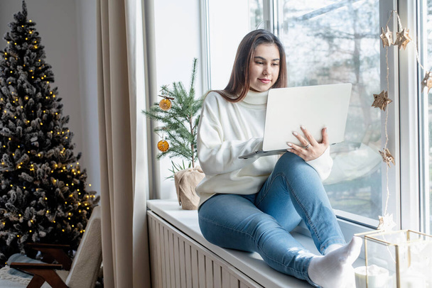 Merry Christmas and Happy New Year.Woman in warm white winter sweater sitting at windowsill at home at christmas eve working on laptop, fir tree behind - Fotoğraf, Görsel