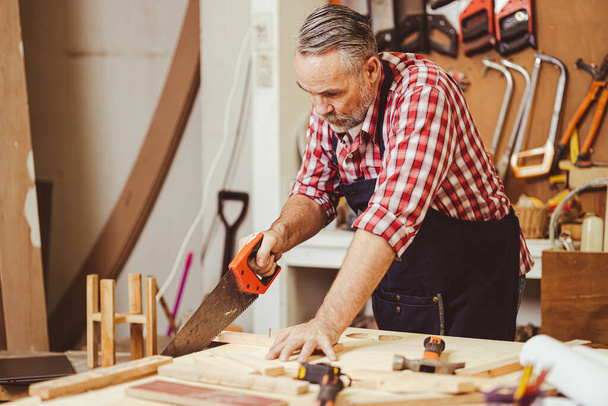 Senior professional male caucasian diy working saw cutting wood making home furniture in wood workshop, home renovate. - Photo, image