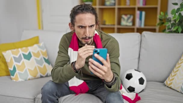 自宅でサポートしているスマートフォンでサッカーゲームを見ている若いヒスパニック男性 - 映像、動画