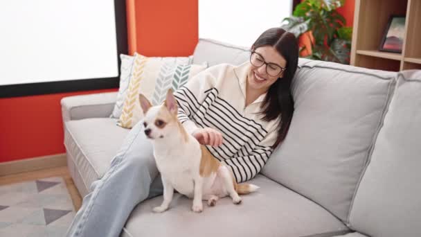 Νεαρή ισπανόφωνη γυναίκα με chihuahua σκύλο χρησιμοποιώντας φορητό υπολογιστή κάθεται στον καναπέ παίζοντας μαζί στο σπίτι - Πλάνα, βίντεο