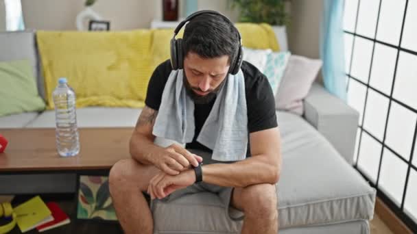Jovem hispânico sentado no sofá ouvindo música tocando relógio em casa - Filmagem, Vídeo