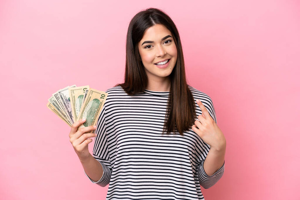 Young Brazilian woman taking a lot of money isolated on pink background giving a thumbs up gesture - Фото, изображение