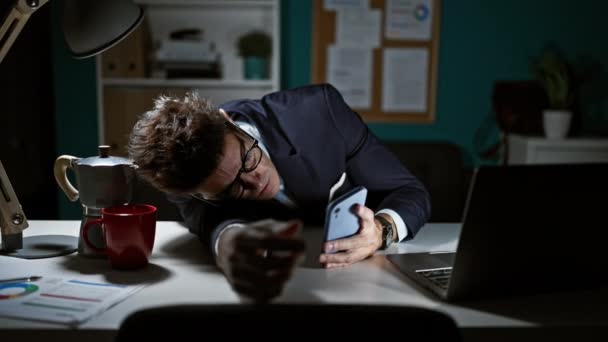 若いヒスパニックの男のビジネスマンは,オフィスでスマートフォンを使用してテーブルの上に疲れています - 映像、動画