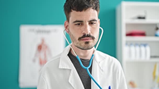 Młody latynoski lekarz uśmiecha się pewnie badając stetoskopem w klinice - Materiał filmowy, wideo