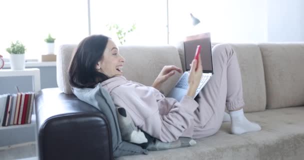 Een vrouw die op een bank ligt met een laptop neemt een videogesprek aan op een smartphone. Vreugdevolle begroeting per telefoon, internet relatie - Video