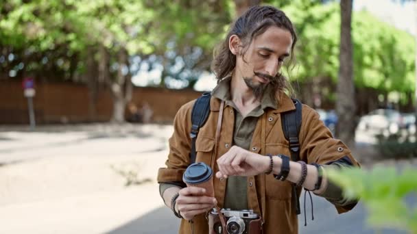 Young hispanic man tourist looking watch holding coffee at park - Footage, Video