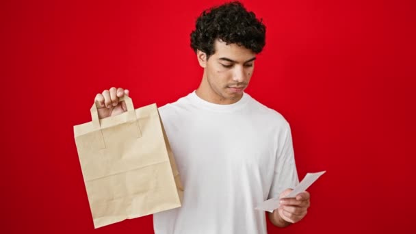Mladý latinský muž drží papírové tašky a lístek přes izolované červené pozadí - Záběry, video