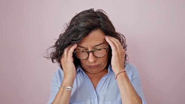 Hiszpanka w średnim wieku zdejmująca okulary cierpiąca na ból głowy na odizolowanym różowym tle - Materiał filmowy, wideo