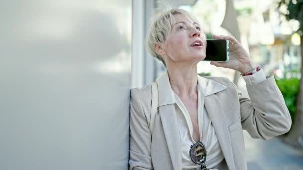 Blondynka w średnim wieku uśmiechnięta pewnie rozmawiająca na smartfonie na ulicy - Materiał filmowy, wideo