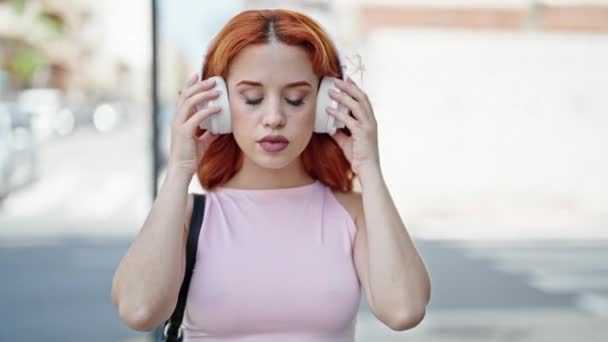 Νεαρή κοκκινομάλλα γυναίκα χαμογελά αυτοπεποίθηση λαμβάνοντας έξω ακουστικά στο δρόμο - Πλάνα, βίντεο