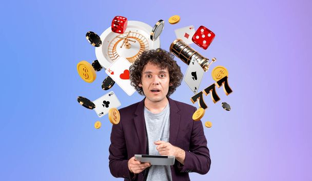 Astonished man finger point at tablet, looking at the camera with mouth opened. Casino jackpot and slot machine icons on gradient background. Concept of big win, luck and gambling - Photo, Image