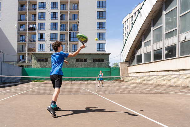 Боковой вид подростка, бьющего теннисный мяч ракеткой, стоя на корте с тенью и играя с анонимной девушкой против жилого дома в солнечный день - Фото, изображение