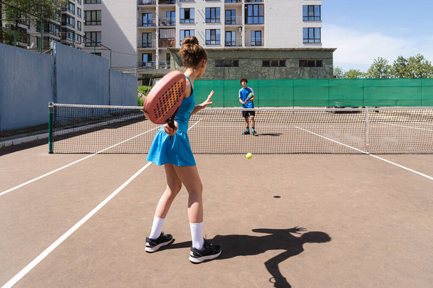 Обратный вид на неузнаваемую молодую девушку, бьющую теннисный мяч ракеткой, стоя на корте с тенью и играя с анонимным подростком возле жилых домов в солнечный день - Фото, изображение
