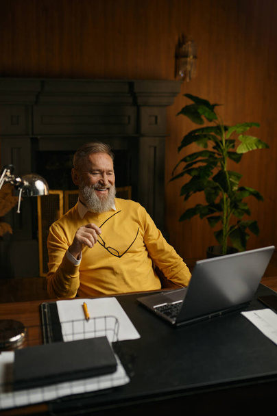 Homme âgé souriant regardant écran d'ordinateur portable pendant le travail ou se reposer au bureau à la maison le soir. Entrepreneur masculin âgé tenant une conférence d'affaires ou regardant une vidéo amusante sur ordinateur - Photo, image