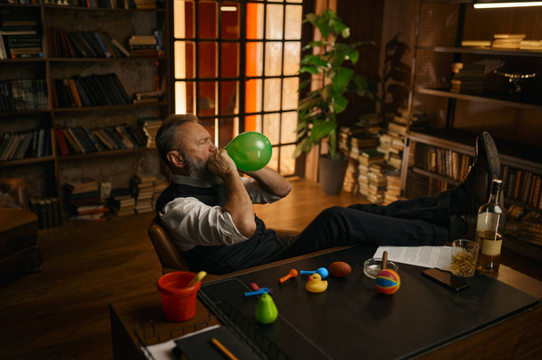 Χαλαρός ανώτερος επιχειρηματίας φορώντας κομψά ρούχα ανατινάζει μπαλόνι ενώ κάθεται στο γραφείο στο σπίτι - Φωτογραφία, εικόνα