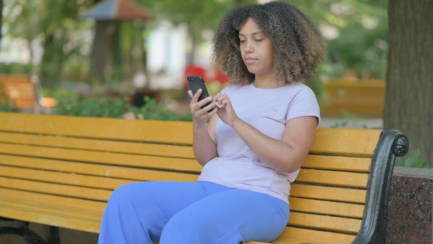 jong Afrikaans vrouw met behulp van smartphone terwijl zitten buiten op een bank - Foto, afbeelding