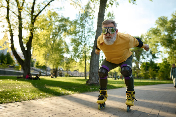 Senior man wearing funny goggles enjoying speed ride on roller skates over city park background at summer day - Photo, Image