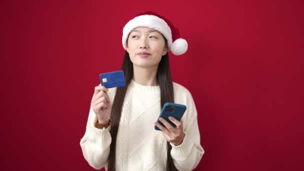 Fiatal kínai nő visel karácsonyi kalap segítségével okostelefon és hitelkártya izolált piros háttér - Felvétel, videó