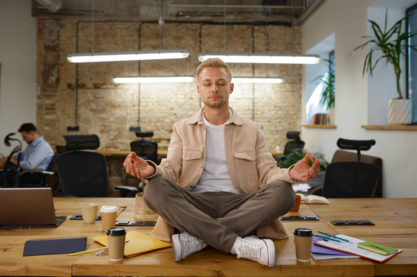 Calm man office worker meditating and breathing sitting in lotus position on desk table at workplace - Photo, Image