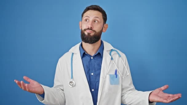 młody Latynos człowiek lekarz stojąc bez pojęcia na odizolowanym niebieskim tle - Materiał filmowy, wideo