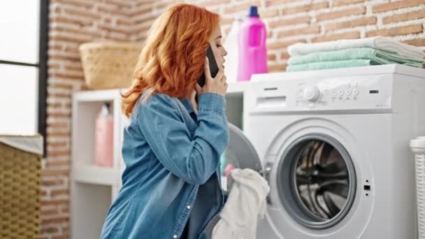Młoda ruda kobieta mówi na smartfonie pranie ubrania w pralni - Materiał filmowy, wideo