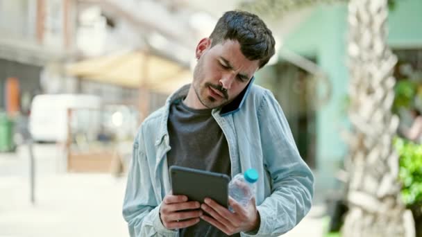 Jonge Spaanse man met behulp van touchpad praten op smartphone houden fles water op straat - Video