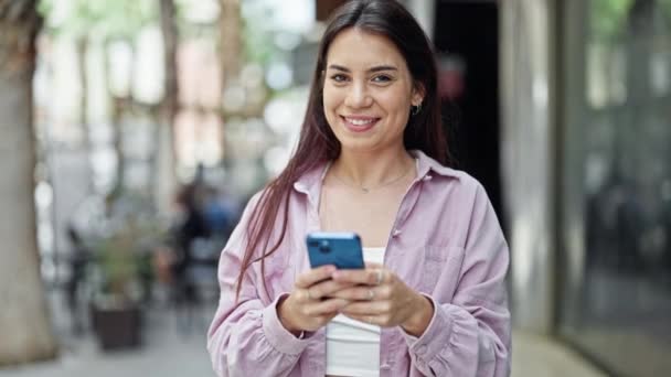 Νέα όμορφη ισπανόφωνη γυναίκα χαμογελά αυτοπεποίθηση χρησιμοποιώντας smartphone στο δρόμο - Πλάνα, βίντεο