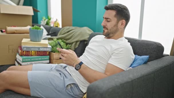 Hombre hispano joven usando teléfono inteligente sentado en un sofá en un nuevo hogar - Metraje, vídeo