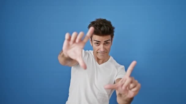Joven hombre hispano haciendo gesto fotográfico con las manos sonriendo sobre un fondo azul aislado - Metraje, vídeo