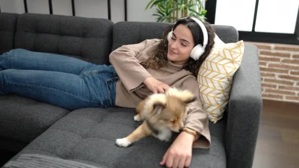 Młoda Latynoska kobieta z psem słuchającym muzyki leżącej na kanapie w domu - Materiał filmowy, wideo