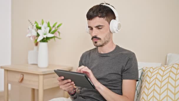 Nuori latino mies käyttää kosketuslevy ja kuulokkeet istuu sängyllä makuuhuoneessa - Materiaali, video
