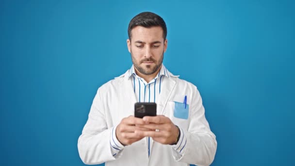 Giovane ispanico uomo medico sorridente utilizzando smartphone su isolato sfondo blu - Filmati, video