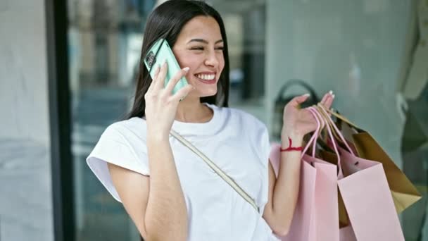 Jovem bela mulher hispânica falando no smartphone segurando sacos de compras na loja de roupas - Filmagem, Vídeo