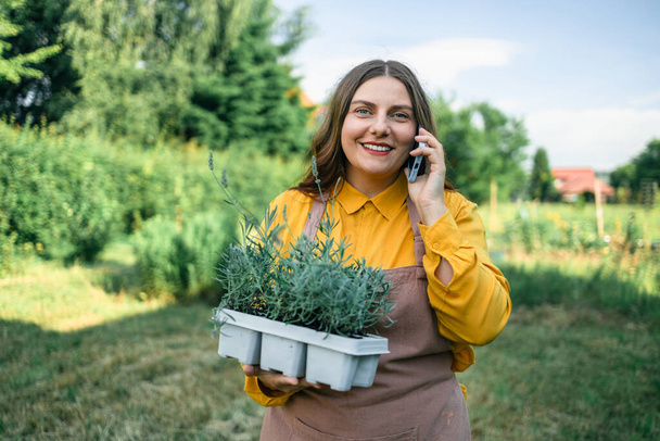Tiro de um agricultor caucasiano fêmea feliz falando no smartphone e segurando um pote de plástico de plantas de flores de lavanda para plantar no chão. Jardineiro usando telefone celular  - Foto, Imagem