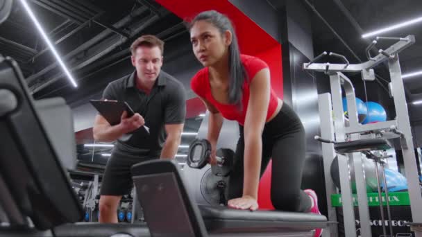 Caucasian man trainer guidance and asian woman lifting dumbbell with motivation and dedication in fitness gym, woman workout for muscular hands and arms with coach support, strength and health. - Footage, Video
