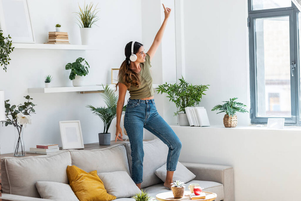 Shot of motivated happy woman singing and dancing on couch while listening music with headphones at home. - Foto, afbeelding