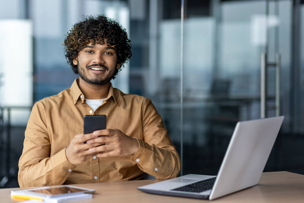 Retrato de um jovem empresário árabe bem sucedido dentro do escritório, o homem está sorrindo e olhando para a câmera, o funcionário está usando um aplicativo no telefone, navegando páginas on-line - Foto, Imagem