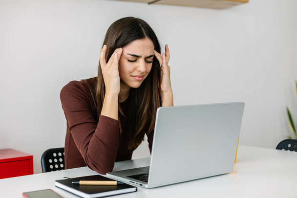 Νεαρή ενήλικη γυναίκα που πάσχει από πονοκέφαλο ή ημικρανία λόγω της εξέτασης της οθόνης laptop ενώ εργάζεται στο σπίτι. - Φωτογραφία, εικόνα