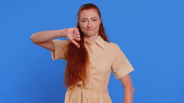 Niechęć. Zdenerwowana nieszczęśliwa młoda kobieta pokazuje kciuki w dół znak gest, wyrażając niezadowolenie, dezaprobatę, niezadowolenie, niechęć. dość atrakcyjne ruda dziewczyna odizolowany na niebieski studio tło - Materiał filmowy, wideo