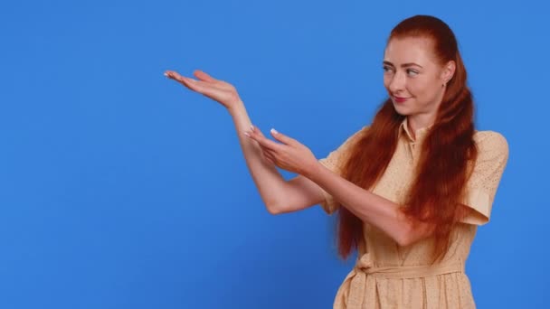 Young woman showing thumbs up and pointing empty place, advertising area for commercial text, copy space for goods promotion advertisement. Redhead girl isolated on blue studio background. Lifestyles - Footage, Video