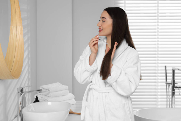 Happy young woman applying essential oil onto hair near mirror in bathroom - Photo, Image