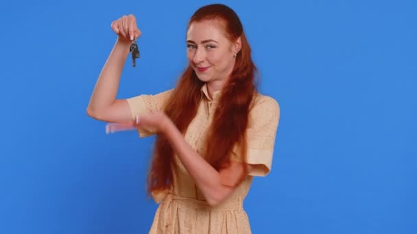 Young adult woman real estate agent lifting hand showing the keys of new home house apartment, buying or renting property, mortgage loan. Redhead pretty girl isolated on blue studio background indoors - Footage, Video