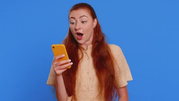 Νεαρή ενήλικη γυναίκα που αναζητούν οθόνη smartphone ειλικρινά χαρά νίκη, λαμβάνοντας χρήματα δολάρια μετρητά χαρτονομίσματα, επιτυχία τύχη λοταρία, γιορτάζει. Redhead κορίτσι απομονωμένο σε μπλε φόντο στούντιο - Πλάνα, βίντεο
