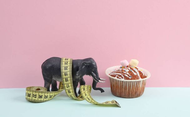 Zabawkowy słoń z babeczką i taśmą pomiarową na różowym niebieskim tle. Dieta, koncepcja odchudzania - Zdjęcie, obraz